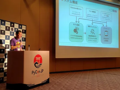 PyCon JP 2026 の会場で、システム構成図を映したスクリーンの横に立ち、演台でマイクを持って話す寺田学